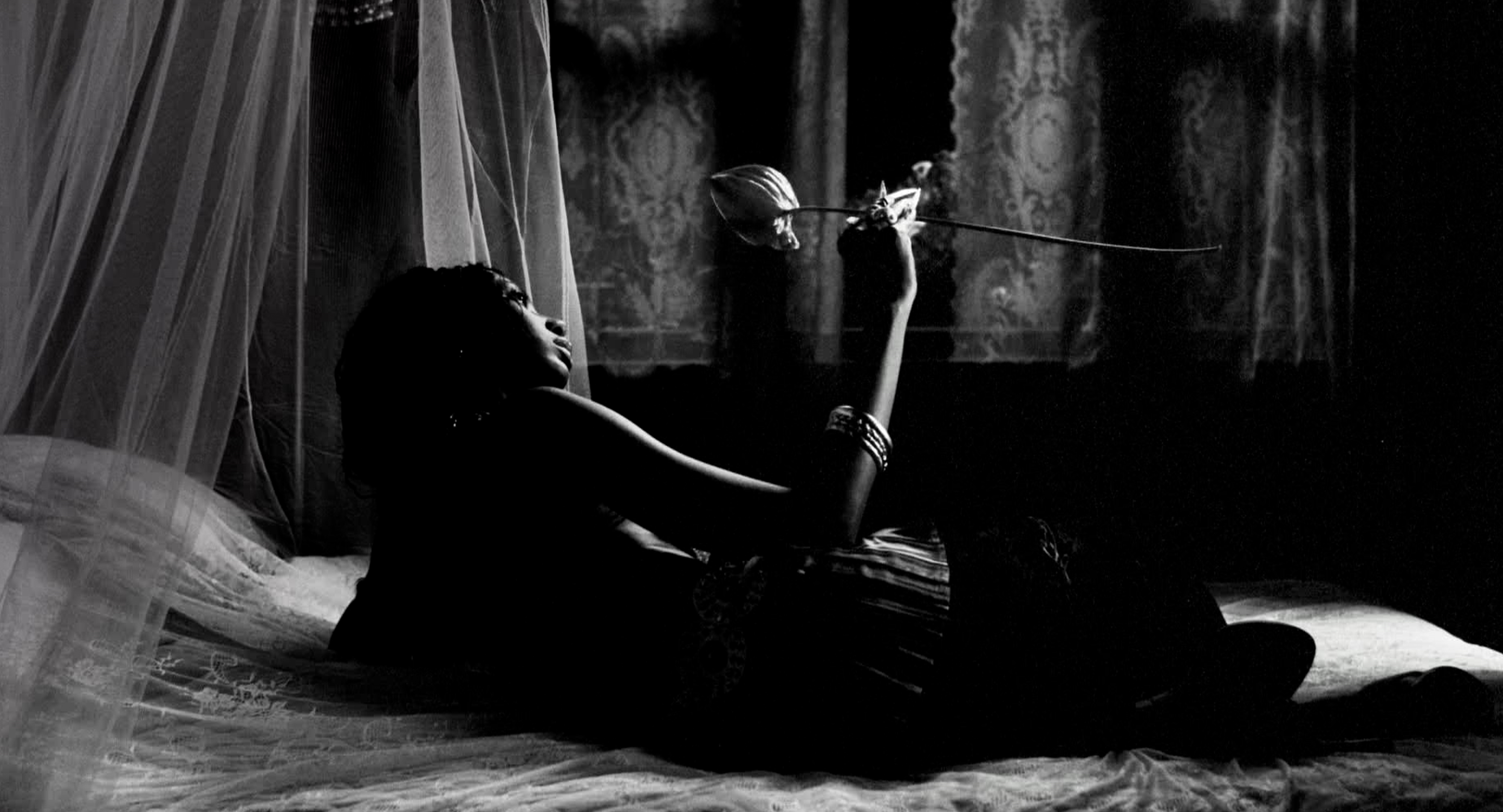 Black and white shot of a model with deep skin looking into a flower while reclining on a bed